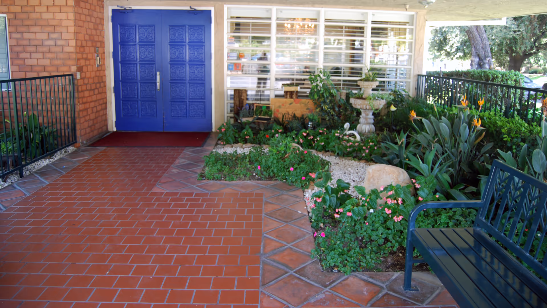 Entrance area of a building with a blue double door, a tiled floor, a garden with various plants and flowers, a stone fountain, and a green bench on the right side.