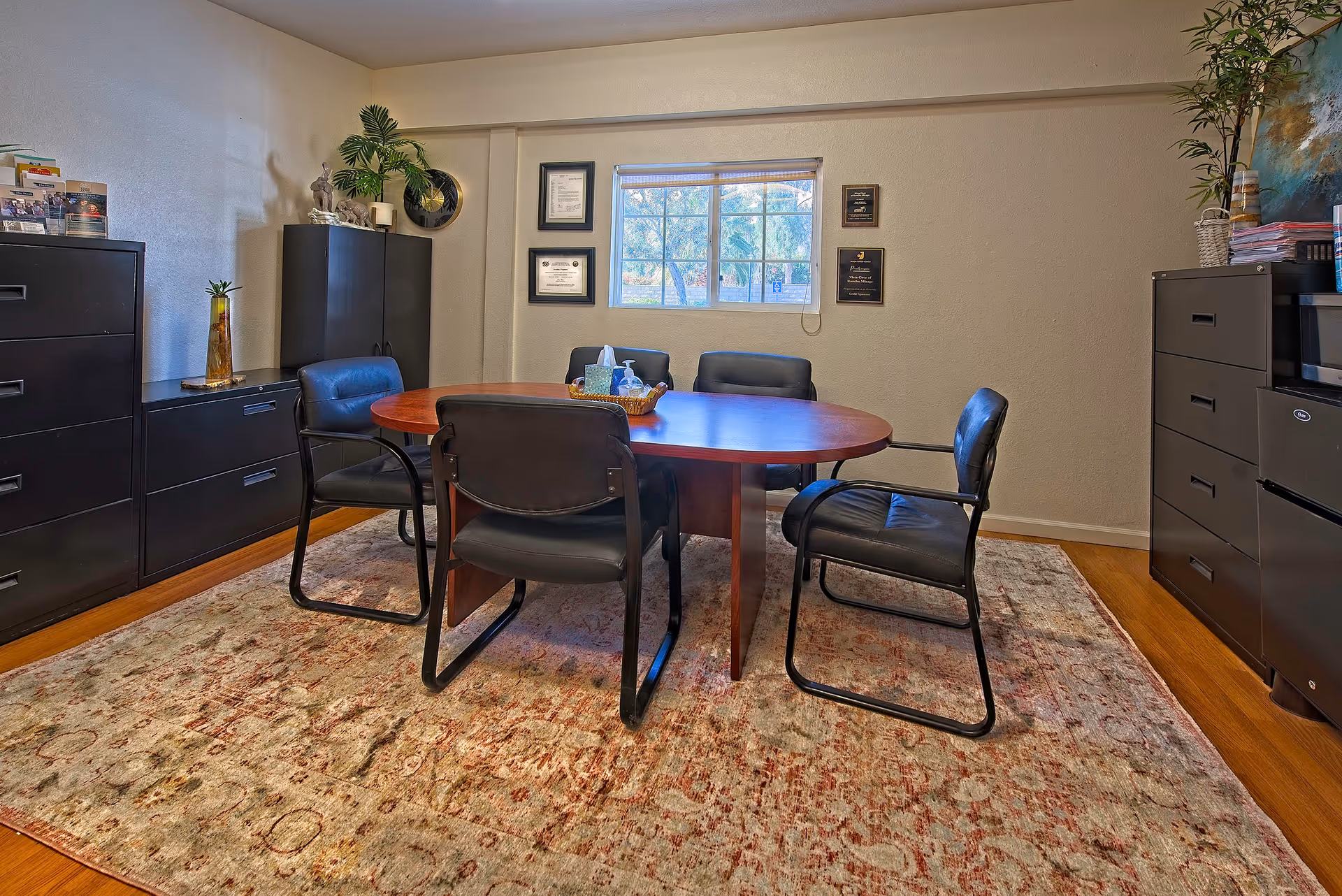 Small office meeting room with an oval wooden table surrounded by black chairs, filing cabinets, a window, and framed certificates on the wall.