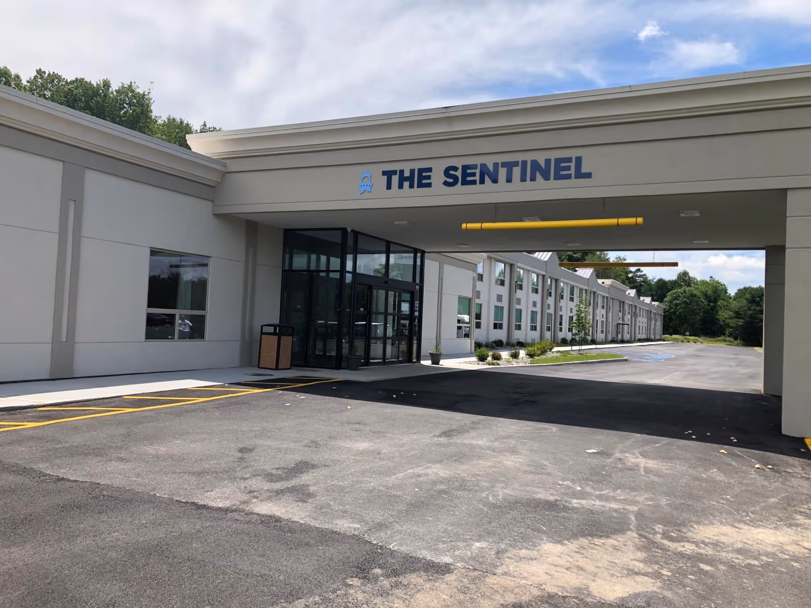 Exterior view of The Sentinel senior living facility showing the main entrance with a covered driveway, glass doors, and a long building with multiple windows extending to the right. The area is paved with parking spaces and surrounded by greenery and trees.