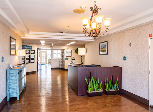 Reception area of Park Regency Retirement Center with a wooden floor, a dark wood reception desk adorned with two potted plants, a chandelier hanging from the ceiling, framed pictures on the walls, a turquoise cabinet with a lamp on the left, and a hallway leading to glass doors in the background.
