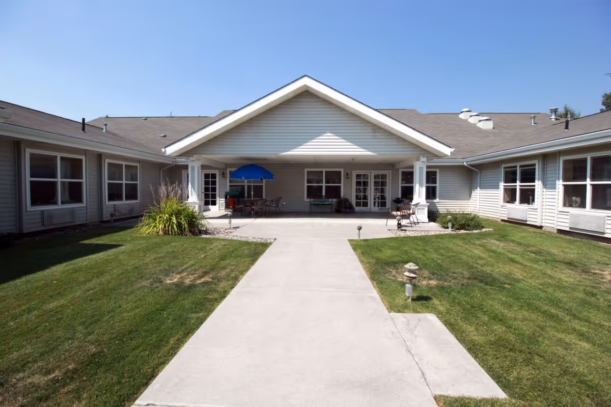 Outdoor view of a senior living facility courtyard with a concrete walkway leading to a covered patio area. The patio has outdoor seating including tables, chairs, and a blue umbrella. The building is single-story with multiple windows and a gray exterior. The lawn is well-maintained with green grass and small garden areas.