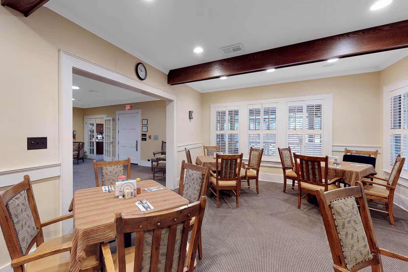 A dining room in an assisted living facility with several wooden tables covered with striped tablecloths and wooden chairs with patterned cushions. The room has beige walls, a carpeted floor, white window shutters, and a dark wooden ceiling beam. In the background, there is an open doorway leading to another room with more seating and a white door.