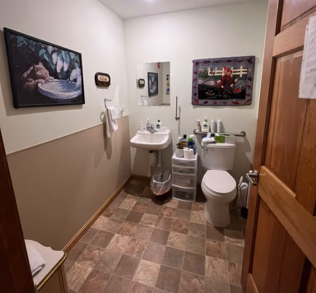 A small bathroom with a white sink and mirror on the wall, a toilet with various toiletries on top and next to it, a towel hanging on a rack, a trash bin, and two framed pictures on the walls. The floor is covered with square tiles in shades of brown and beige.