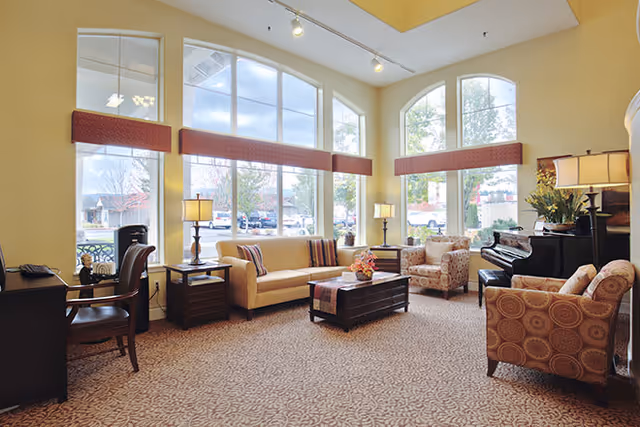 A bright and spacious living room area with large arched windows letting in natural light. The room features a beige sofa, two patterned armchairs, a wooden coffee table with a flower arrangement, two side tables with lamps, a piano, and a desk with a chair. The carpet has a subtle pattern and the walls are painted a light yellow.