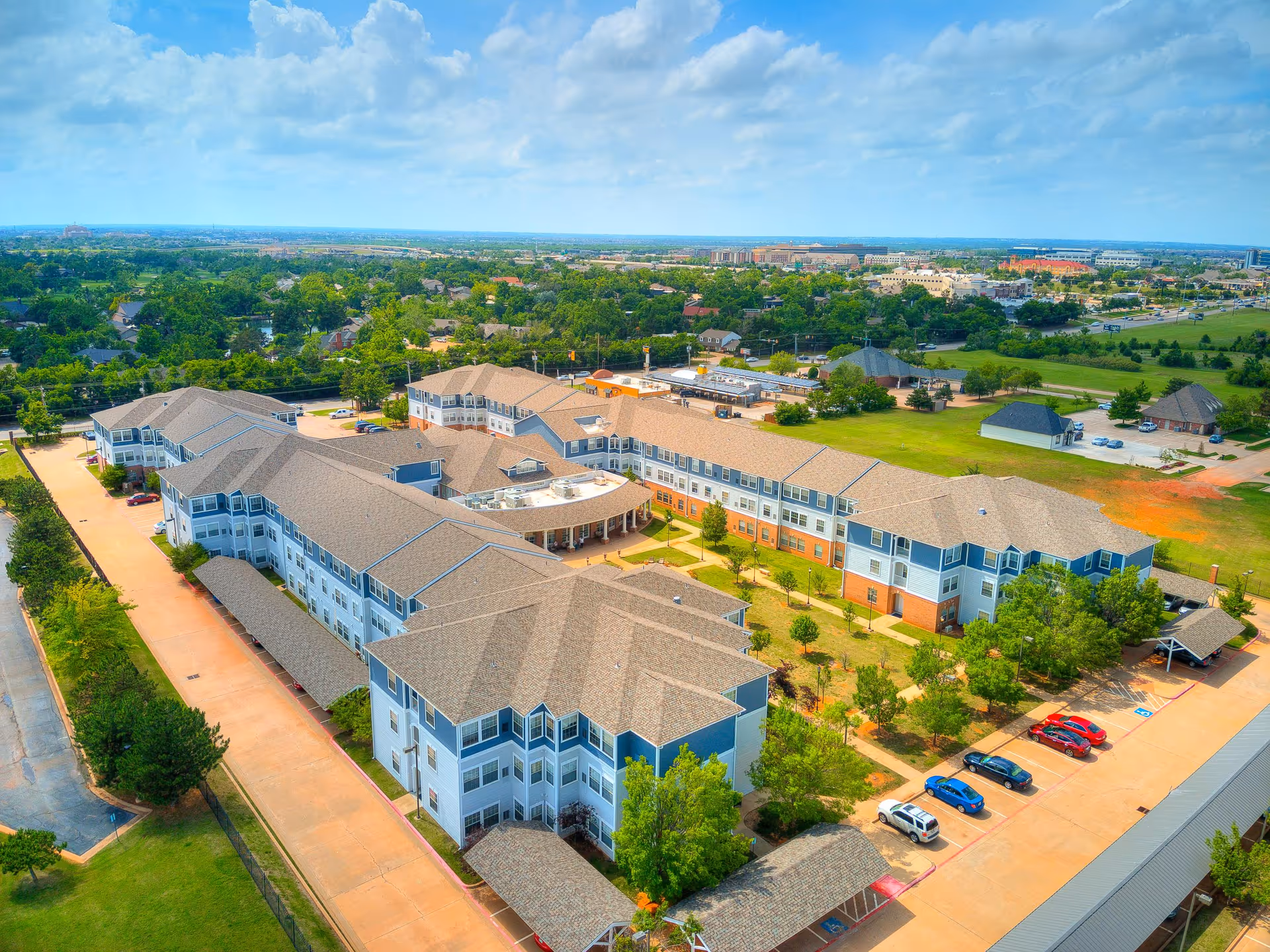 Aerial view of a large senior living facility named Town Village, featuring multiple connected buildings with beige roofs and blue-gray walls, surrounded by green lawns, trees, and parking areas with several cars. The facility is situated in a suburban area with other buildings and greenery visible in the background under a partly cloudy sky.
