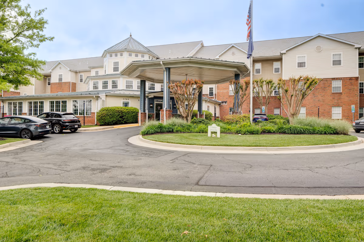 Front exterior view of Arbor Terrace Sudley Manor, a multi-story senior living facility with a covered entrance, landscaped greenery, and several parked cars in front.