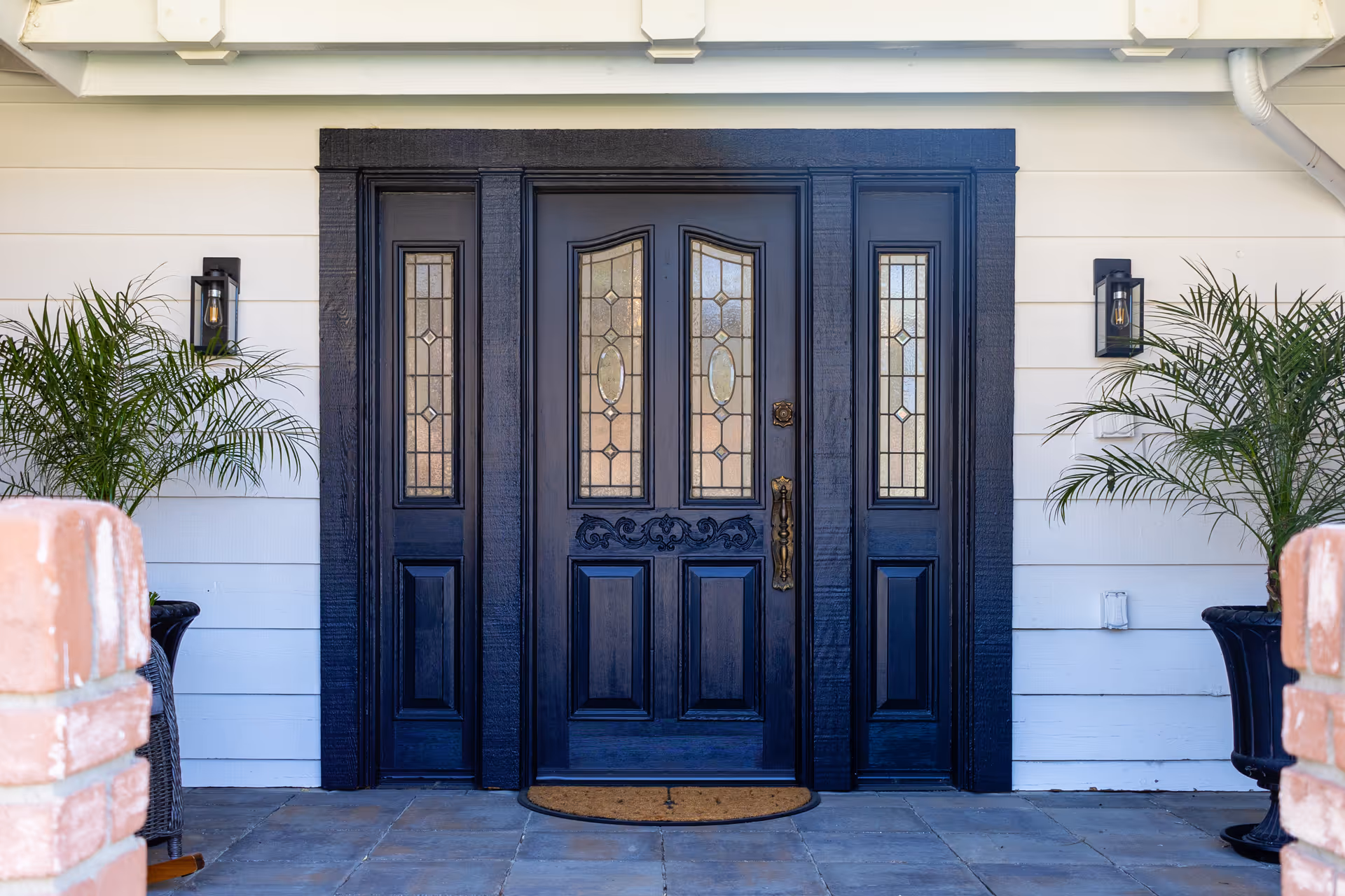 Front entrance of a building with a black door featuring decorative glass panels, flanked by two potted palm plants and two wall-mounted lantern-style lights on a white exterior wall.