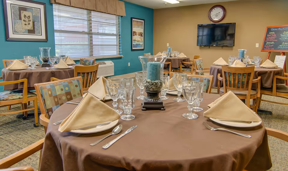 A dining room in a senior living facility with round tables covered in brown tablecloths, each set with beige folded napkins, plates, silverware, and multiple clear glasses. The chairs have patterned cushions. The room has a teal accent wall with framed artwork, a window with blinds, a wall-mounted TV, and a chalkboard menu on the wall.