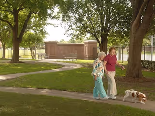Two elderly women walking on a paved path in a green outdoor area with trees and grass. One woman is wearing a floral shirt and blue pants, and the other is wearing a red shirt and beige pants. They are walking a small dog on a leash.