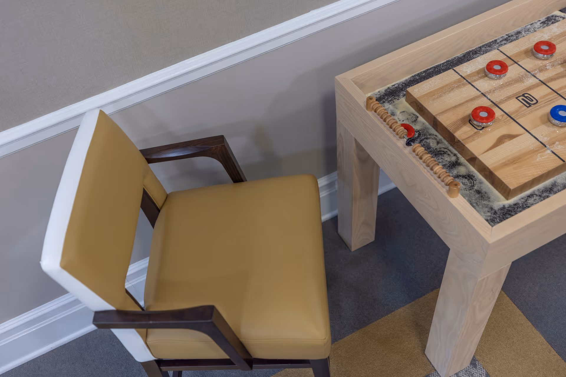 A close-up view of a beige and white cushioned chair with wooden armrests next to a wooden shuffleboard game table with red and blue pucks on a carpeted floor.