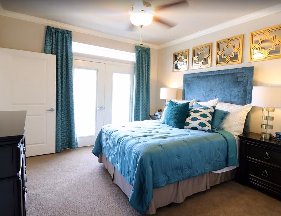 A bright bedroom with a large bed featuring a blue upholstered headboard and teal bedding. The bed is adorned with multiple pillows in shades of teal and white. On either side of the bed are dark wooden nightstands with modern lamps. Above the headboard are four decorative gold-framed mirrors. The room has beige carpet and light-colored walls, with a ceiling fan overhead. French doors with teal curtains allow natural light into the room.