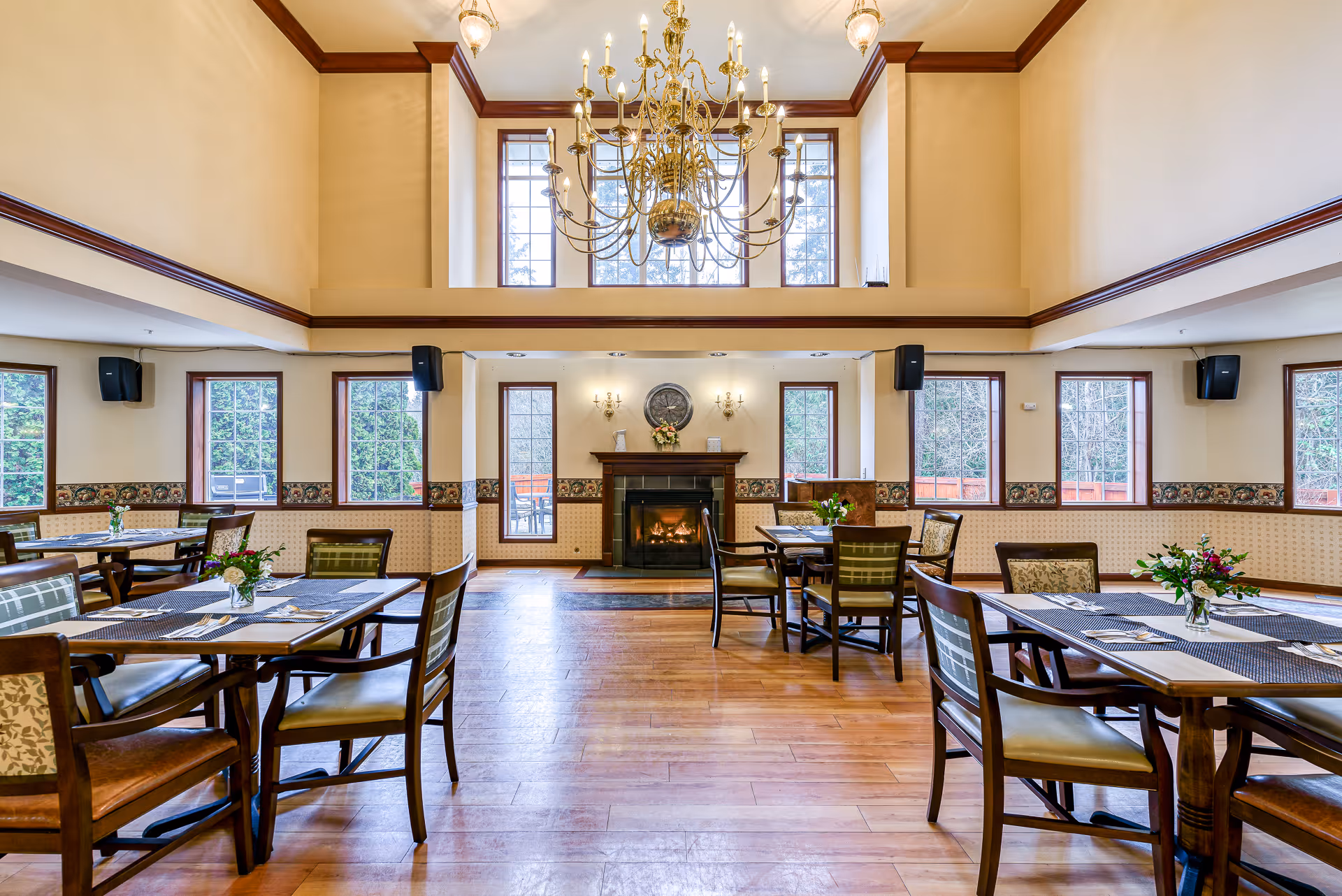 Spacious dining room with wooden tables and chairs, a central chandelier and a fireplace beneath tall windows.