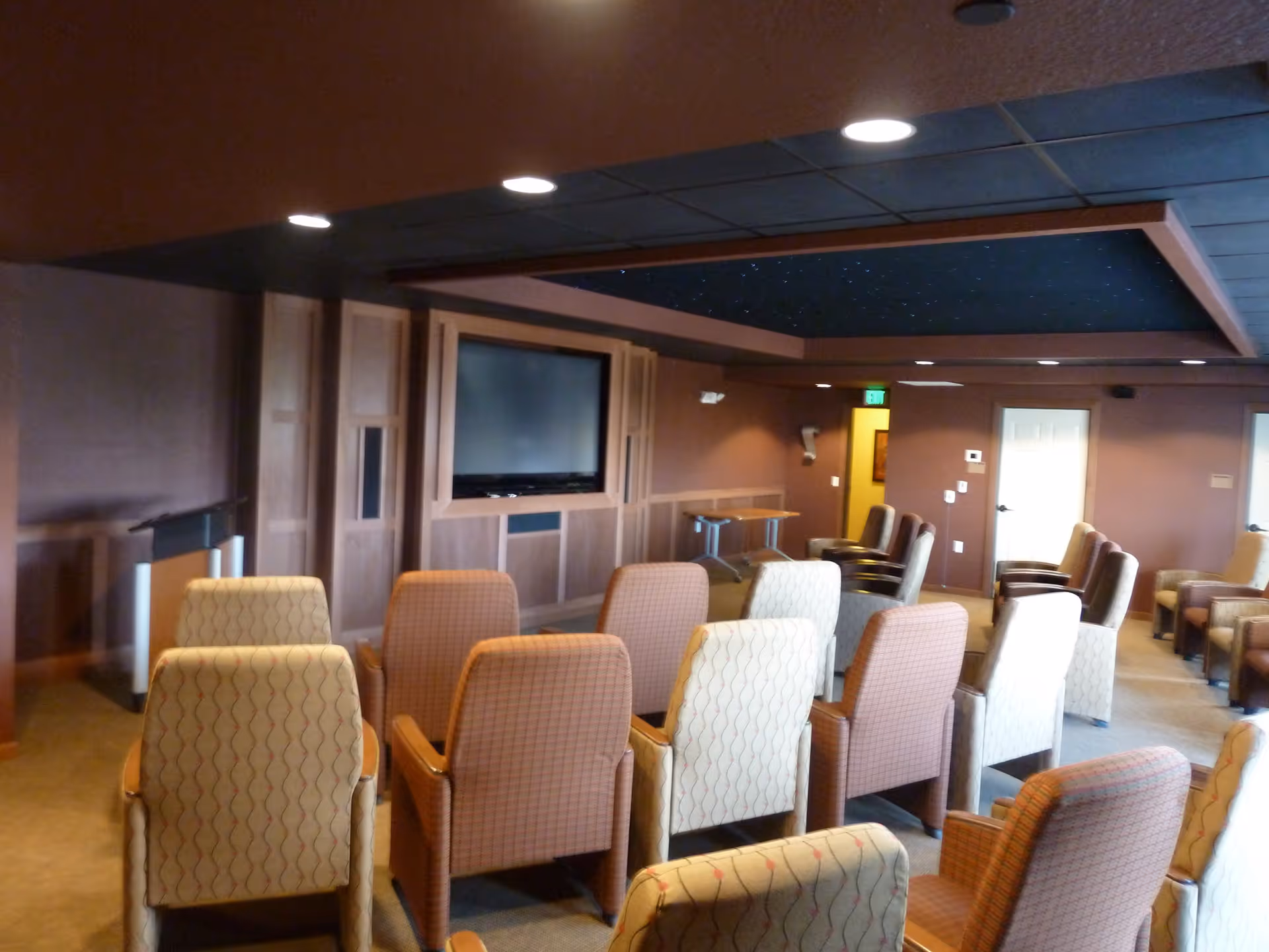 Small theater-style common room with rows of upholstered chairs facing a wall-mounted TV and a podium.