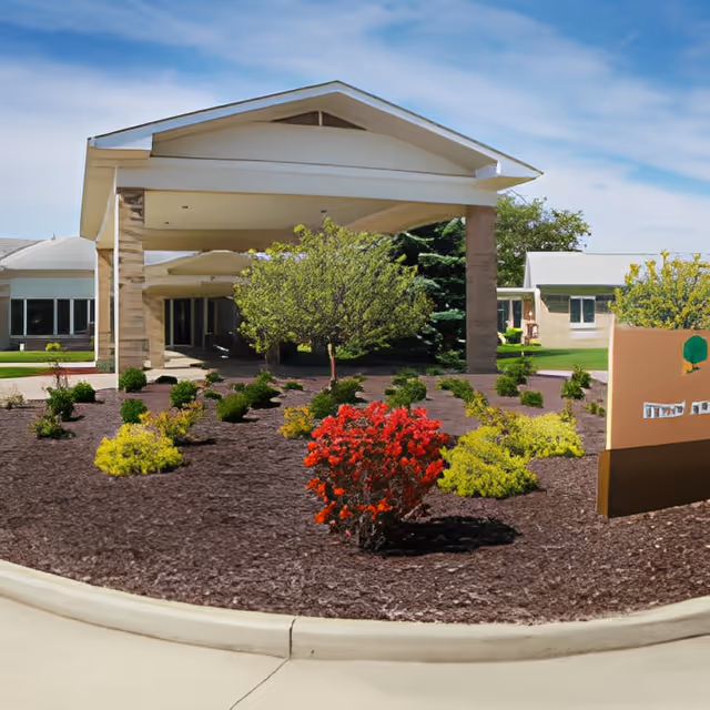 Entrance of Fairlawn Retirement Community featuring a covered drop-off area with stone pillars, landscaped garden with various shrubs and small trees, and a clear blue sky.