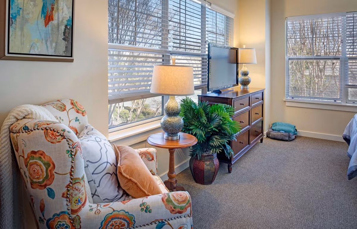 A cozy living area with a floral patterned armchair adorned with two pillows next to a wooden side table holding a decorative lamp. Across from the chair is a wooden dresser with a flat-screen TV and another lamp on top. Large windows with white blinds let in natural light, and a green potted plant is placed beside the dresser. The room has beige walls and carpeted flooring.