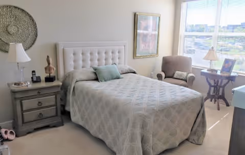 Sunlit bedroom with a tufted headboard bed, bedside tables, an armchair and a large window overlooking the outdoors.