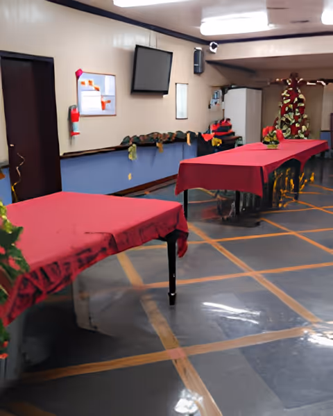 Interior view of a room with two tables covered in red tablecloths, decorated with small green plants. The room has a tiled floor with a grid pattern, beige walls with a blue lower section, a wall-mounted TV, a fire extinguisher, and a bulletin board. There is a white refrigerator and some decorations in the background.