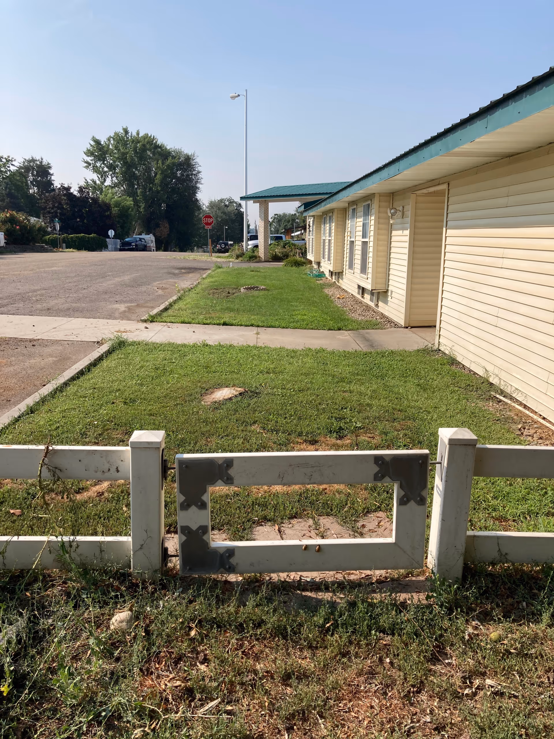 Front lawn with a short white fence leading to a one-story beige building with a green metal roof and a covered entrance and parking area.