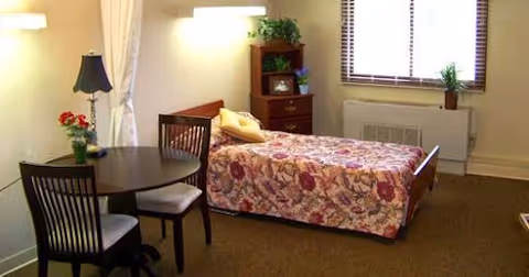 Furnished bedroom with a single bed covered in a floral bedspread, a small round table with two chairs, a dresser, lamp, and a window.