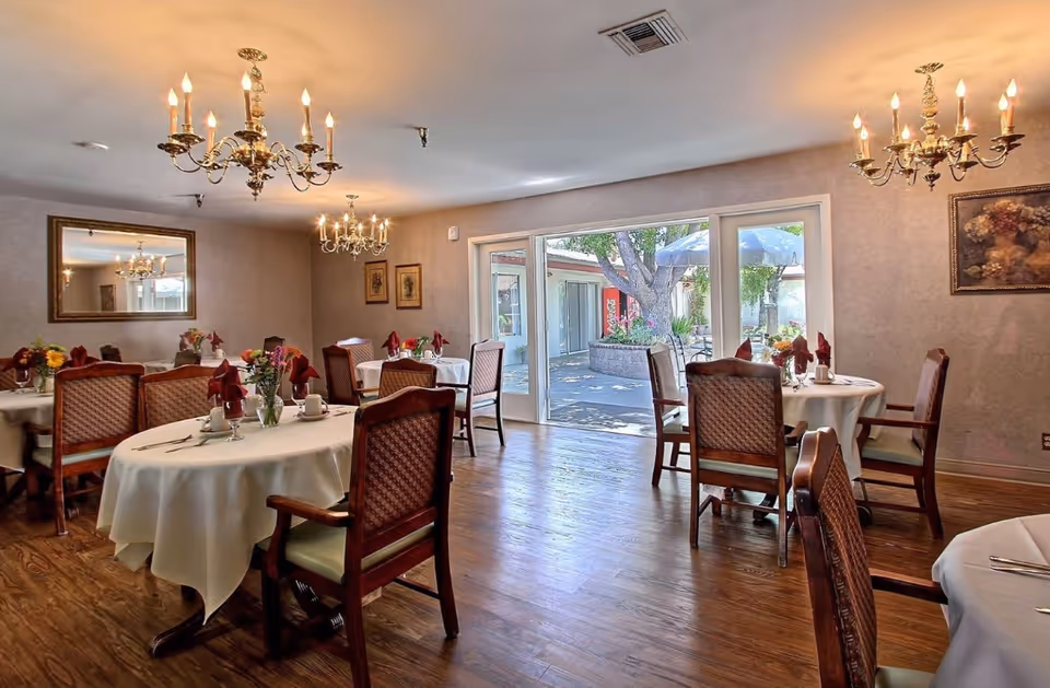 A dining room with round tables covered in white tablecloths, each set with cups, utensils, and red napkins. Wooden chairs with cushioned seats surround the tables. Three ornate chandeliers hang from the ceiling, and a large mirror is mounted on one wall. Glass doors open to an outdoor patio area with trees and umbrellas.