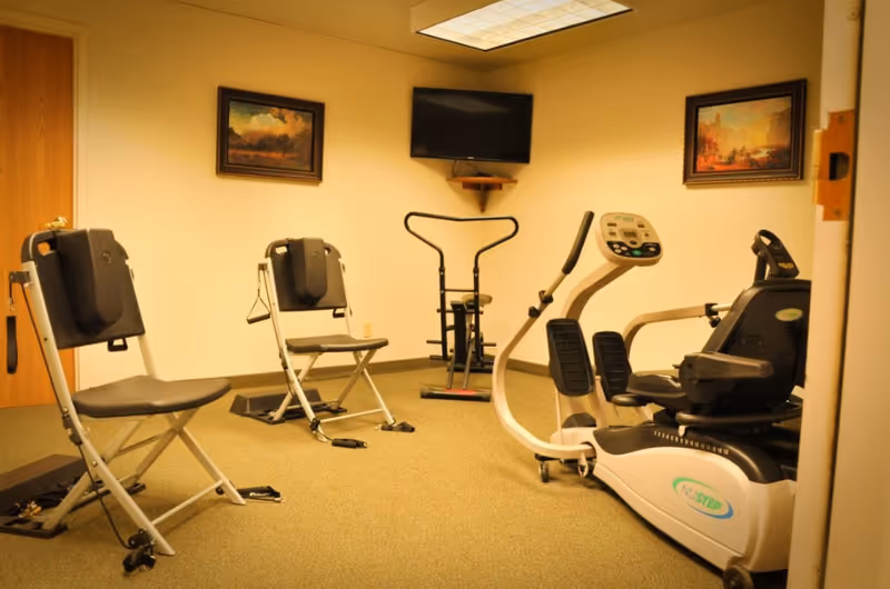Small exercise room with folding exercise chairs, a recumbent cardio machine, a TV mounted on the wall, and framed paintings.