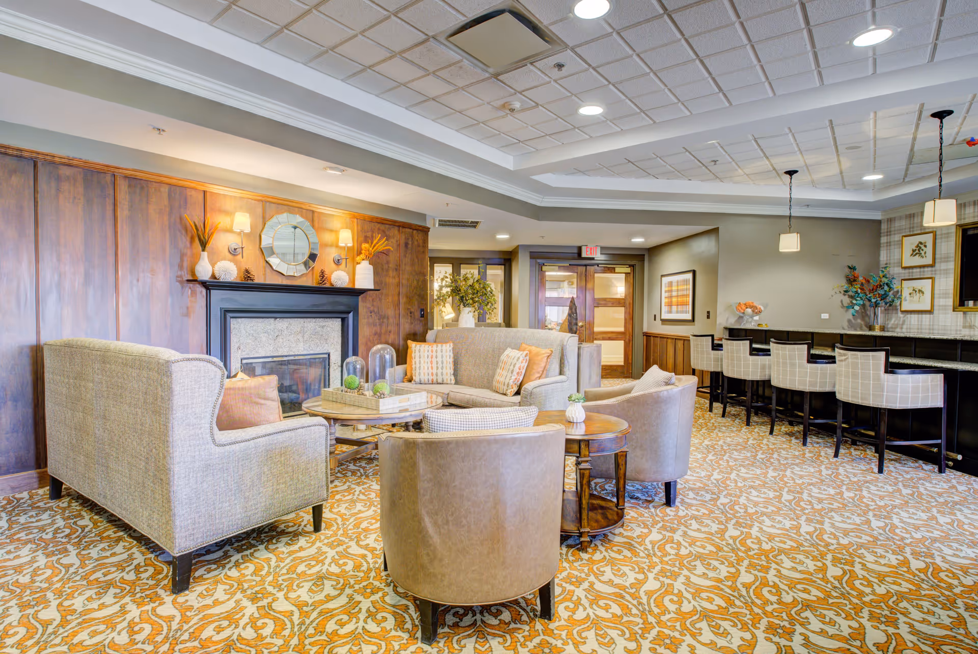 A cozy senior living facility lounge area with beige and brown upholstered chairs and sofas arranged around a wooden coffee table. The room features a decorative fireplace with a round mirror and vases on the mantel, patterned carpet, and a bar area with high chairs. The ceiling has recessed lighting and pendant lights above the bar.