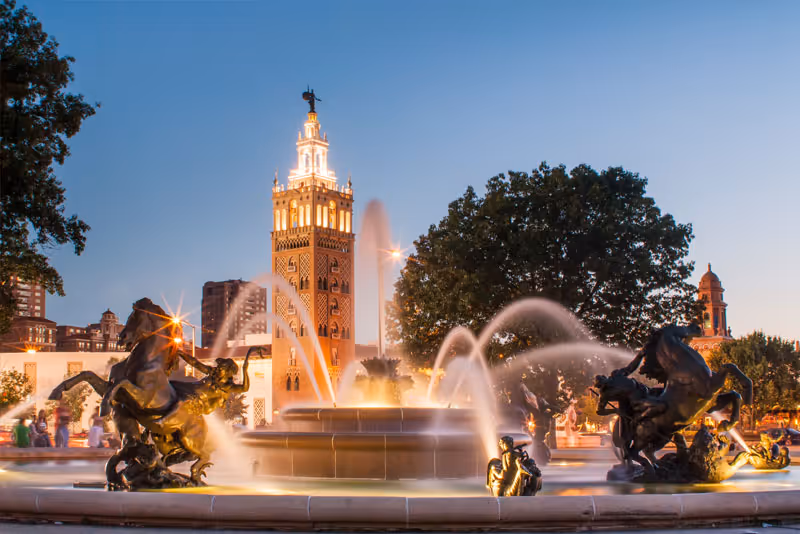 A large decorative fountain with multiple water jets and bronze horse sculptures in an outdoor plaza at dusk, with a tall illuminated tower and trees in the background.
