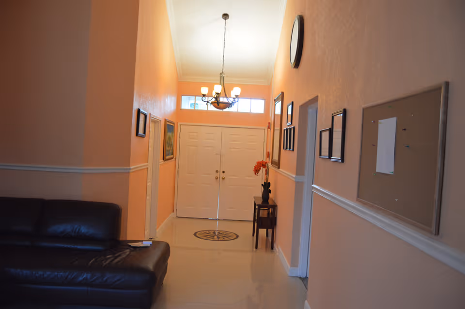 Foyer-style interior with double doors, a chandelier, a black leather sofa, framed pictures and a bulletin board in a peach-colored hallway.