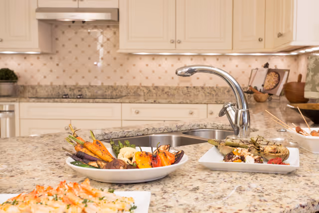 A kitchen countertop with a double sink and chrome faucet. On the granite countertop, there are plates with grilled vegetables and shrimp. The background shows cream-colored cabinets and a tiled backsplash.