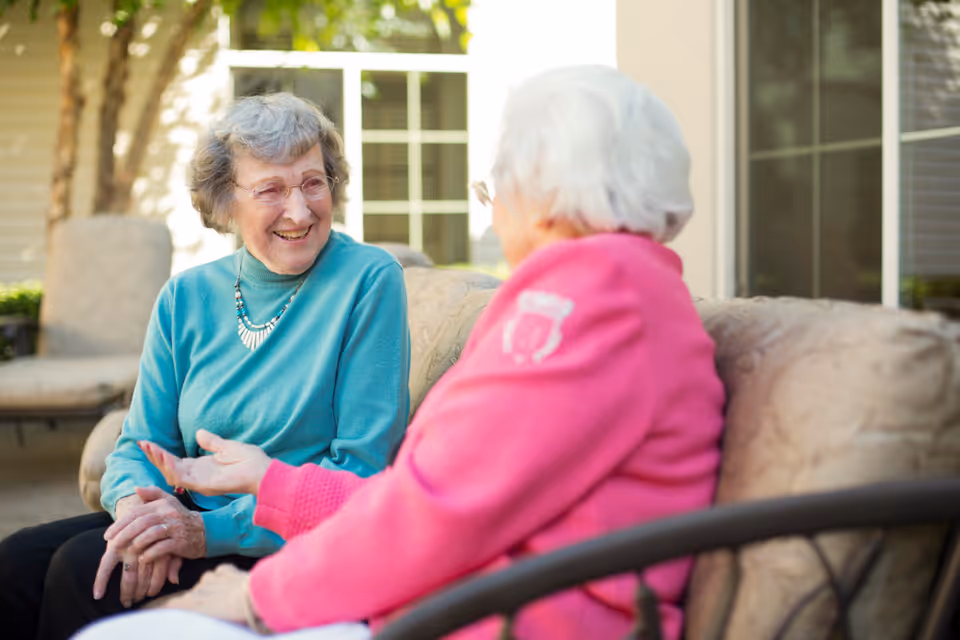 Two elderly women sitting on an outdoor cushioned bench, engaged in a friendly conversation. One woman is wearing a teal sweater and glasses, smiling warmly, while the other woman, with white hair, is wearing a bright pink jacket. The background shows a house exterior with windows and some greenery.