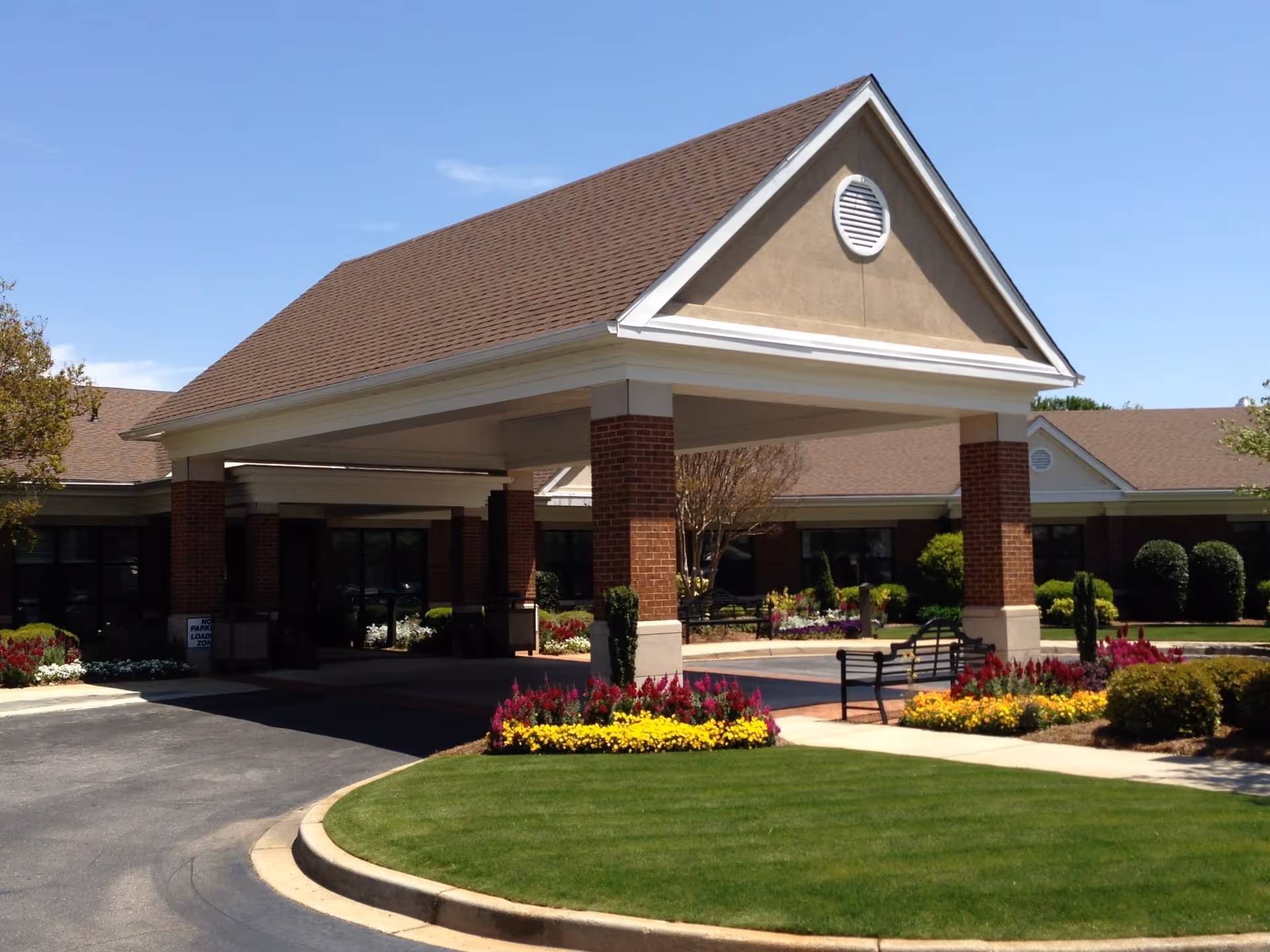 Front entrance of a brick senior living facility with a covered porte-cochere, benches and landscaped flowerbeds.