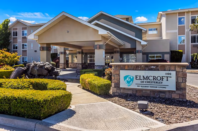 Exterior view of Elmcroft of Chesterley senior living facility showing the entrance with a covered drop-off area, manicured bushes, rock landscaping, and a sign displaying the facility name and logo.