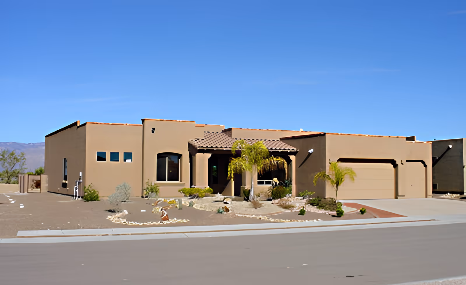 Exterior view of a single-story, beige stucco building with a tiled roof, a covered entrance supported by columns, a two-car garage, and desert landscaping with small shrubs and rocks under a clear blue sky.