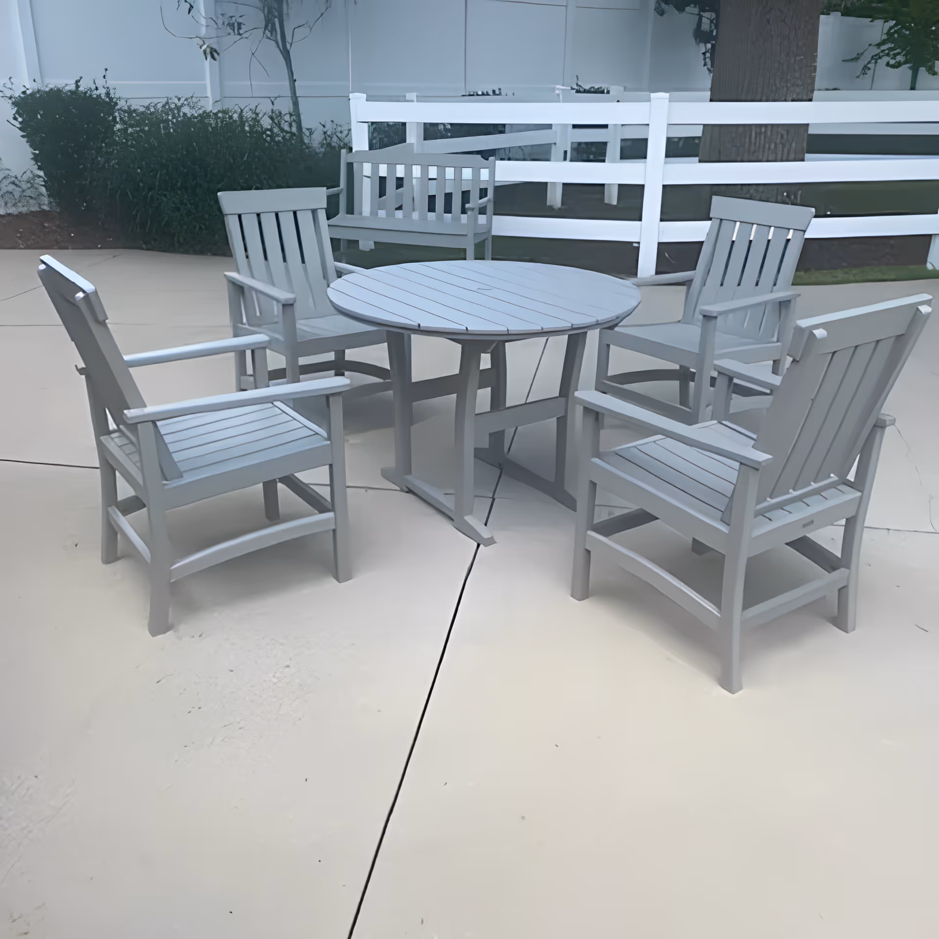 Outdoor patio area with a round table surrounded by four wooden chairs and a wooden bench in the background, all painted in a light gray color. The area is paved and enclosed by a white fence with greenery and a tree visible behind it.