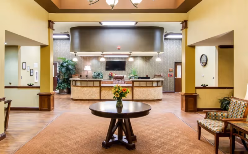 Reception area of Amber Grove Place Memory Care with a round wooden table holding a vase of flowers in the center, a front desk with a chair behind it, a wall-mounted TV, pendant lights, and plants. There are patterned chairs and a clock on the wall to the right.