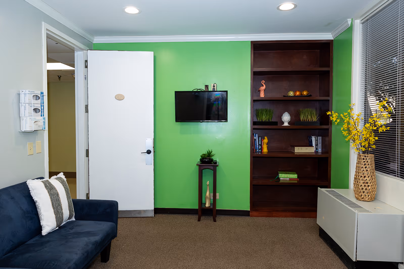 A small lounge area with a dark blue couch with two pillows on the left, a white door slightly open in the center, a wall-mounted flat screen TV on a bright green wall, a small wooden stand with decorative items below the TV, a tall dark wooden bookshelf with various decorations and books on the right, and a window with blinds and a woven vase with yellow flowers on a white radiator cover.