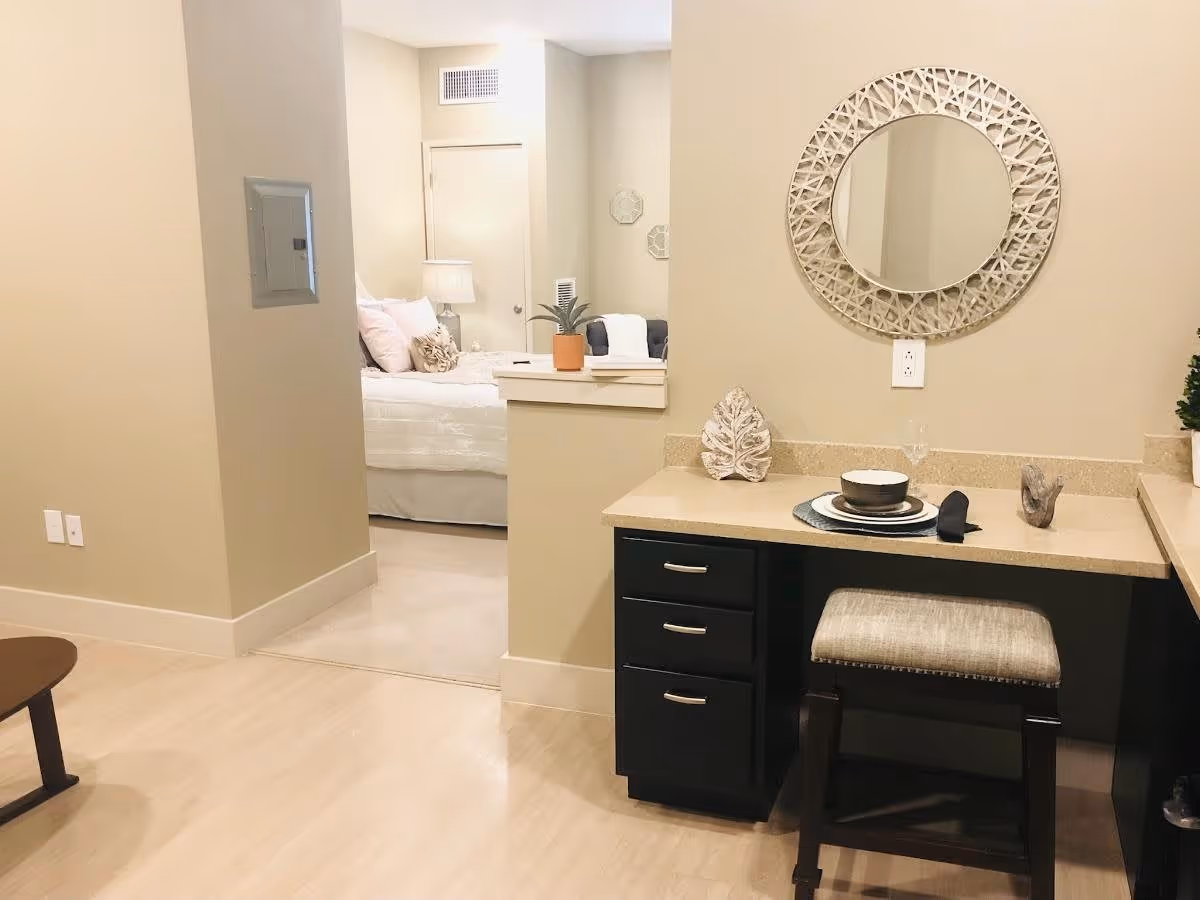 Interior view of a senior living facility showing a small desk area with a round decorative mirror above it, a cushioned stool, and a place setting with a bowl, plate, and napkin. In the background, there is a glimpse of a bedroom with a bed, pillows, a lamp, and a door.