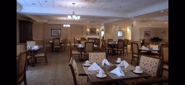 A spacious dining room with multiple tables set with white napkins and cups. The room features cushioned chairs, soft lighting from chandeliers, and framed artwork on the walls. The carpeted floor and neutral color palette create a warm and inviting atmosphere.