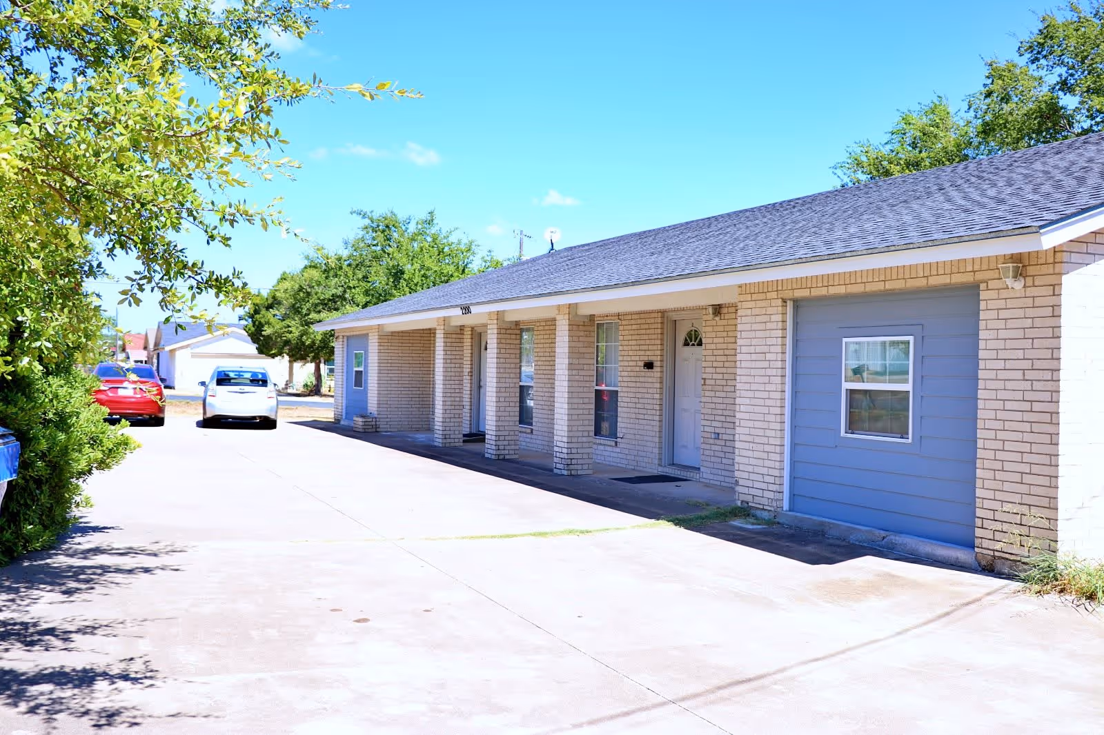 Exterior view of a single-story brick building with a covered porch and a driveway. There are two cars parked on the driveway, and trees and bushes are visible along the side. The sky is clear and blue.