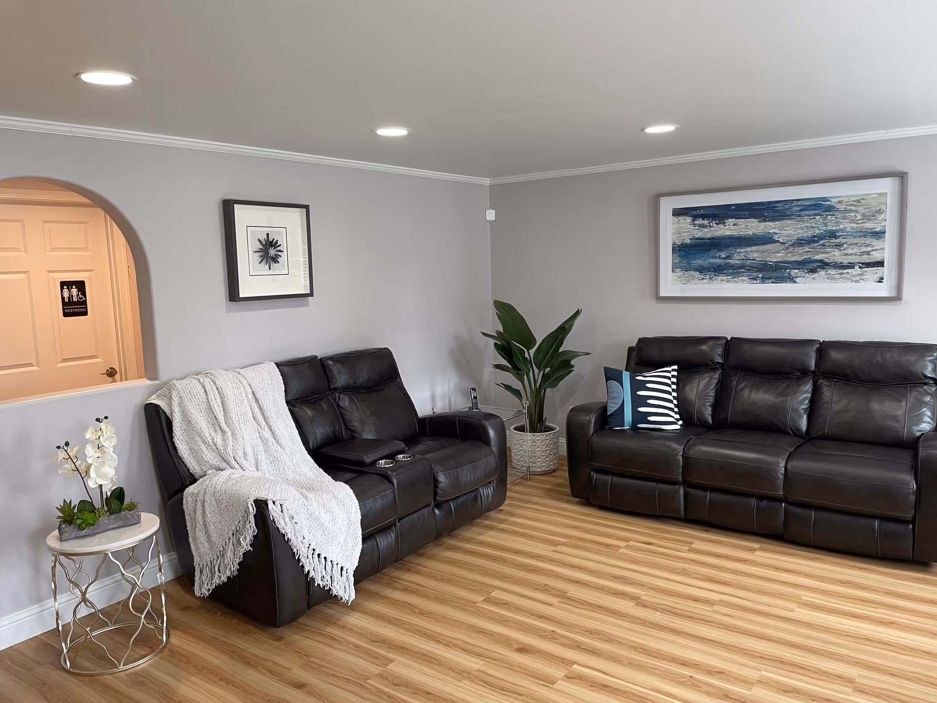 Bright living room with two black leather sofas, artwork on the walls, a potted plant and side table on a wood floor.