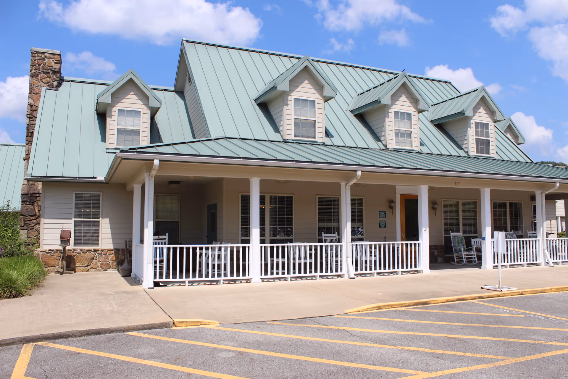Front exterior view of a senior living facility building with a green metal roof, multiple dormer windows, a stone chimney, and a covered porch with white railings and rocking chairs. The sky is blue with some clouds.