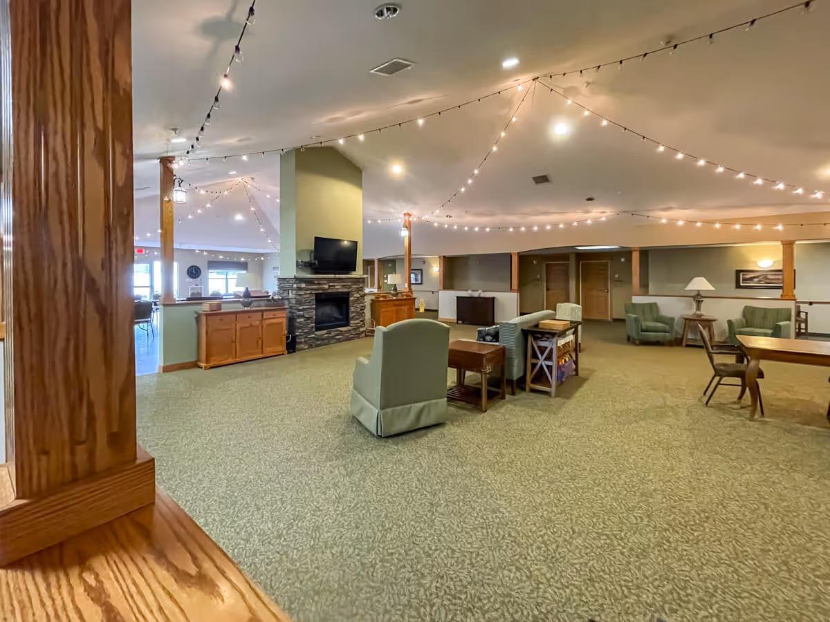 Spacious assisted living common area with green carpet, multiple seating arrangements including armchairs and sofas, wooden tables, a stone fireplace with a mounted TV above it, and string lights hanging from the ceiling creating a warm ambiance.