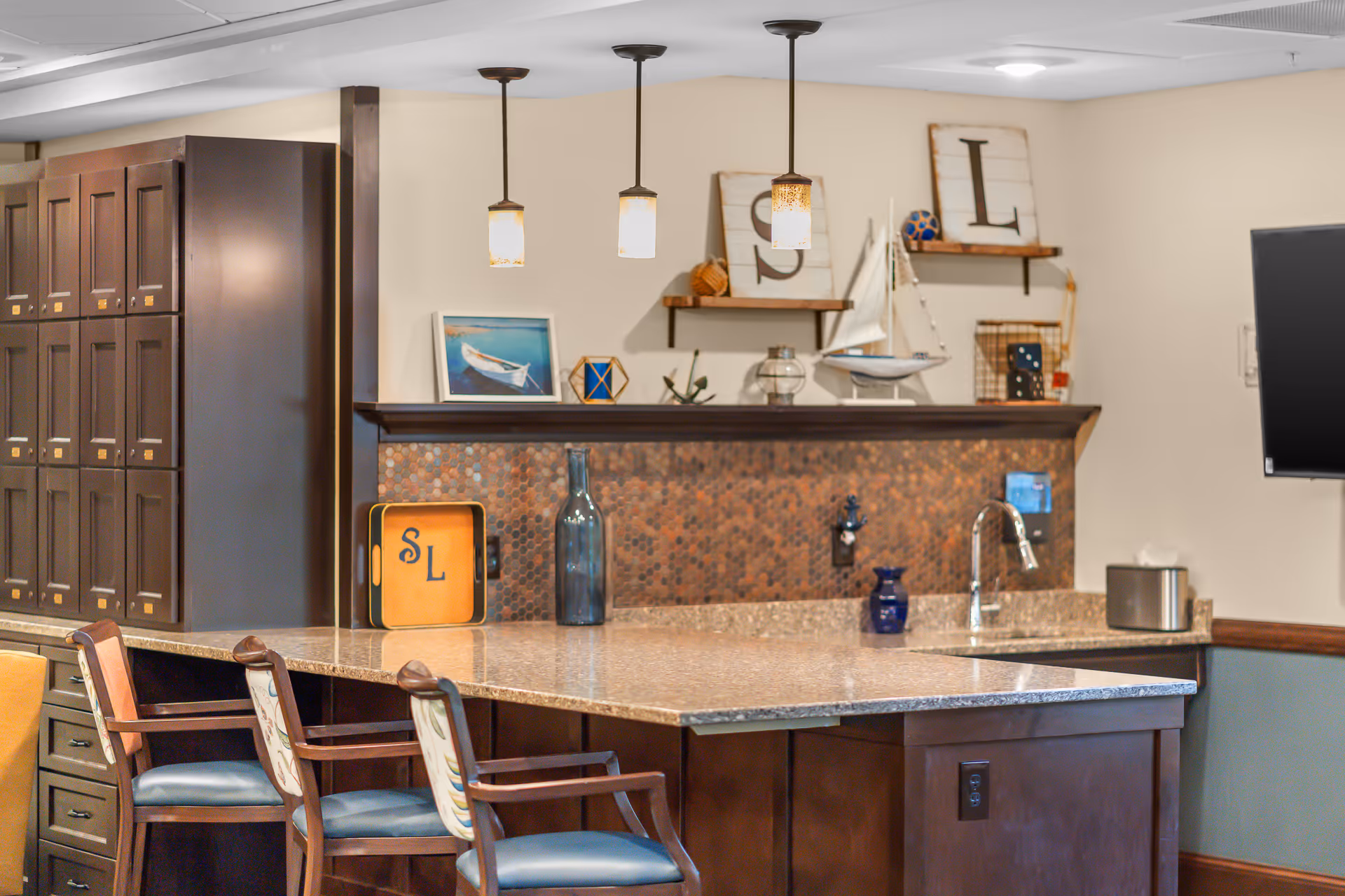 Interior view of a kitchen area with a large granite countertop island surrounded by wooden chairs with blue cushions. The back wall features a copper tile backsplash, a sink with a faucet, and decorative shelves holding nautical-themed items and framed letters 'S' and 'L'. Three pendant lights hang from the ceiling above the island.