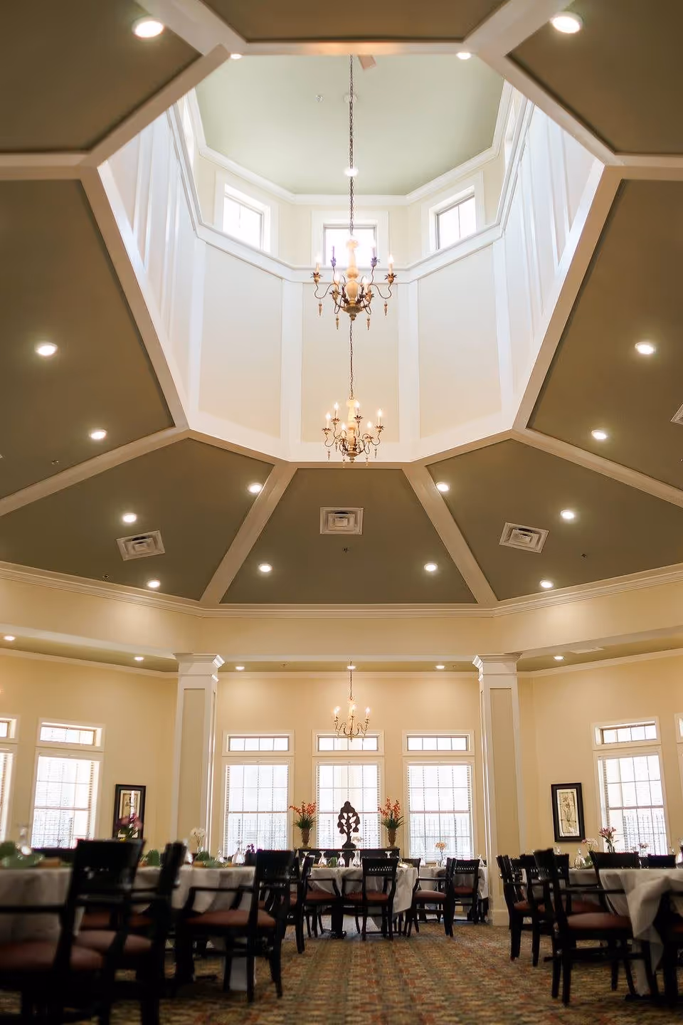 Interior view of a dining room with multiple tables and chairs arranged neatly. The room features large windows allowing natural light to enter, decorative plants on the windowsills, and elegant chandeliers hanging from a high, octagonal ceiling with recessed lighting.
