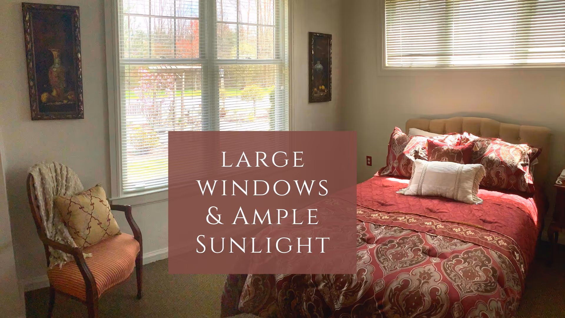 A bedroom with large windows letting in ample sunlight. The room features a bed with a red and gold patterned comforter and multiple pillows. There is a wooden chair with a cushion and a throw blanket next to the window. Two framed paintings hang on the walls beside the windows.