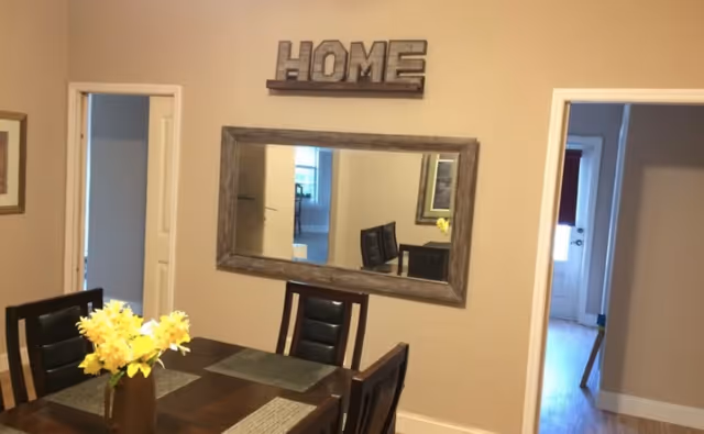 Interior view of a dining area with a wooden table and four chairs. A vase with yellow flowers is placed on the table. On the wall above the table, there is a large rectangular mirror with a wooden frame and a decorative sign that spells 'HOME'. Two doorways are visible on either side of the mirror, leading to other rooms.