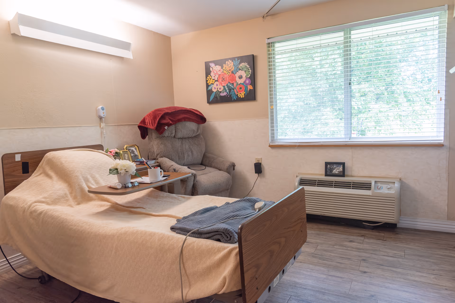 A senior living facility bedroom with a hospital-style bed covered in a beige blanket, a small table with a flower vase and a cup, a gray recliner chair with a red blanket draped over it, a colorful floral painting on the wall, a large window with blinds, and a heating/cooling unit beneath the window.