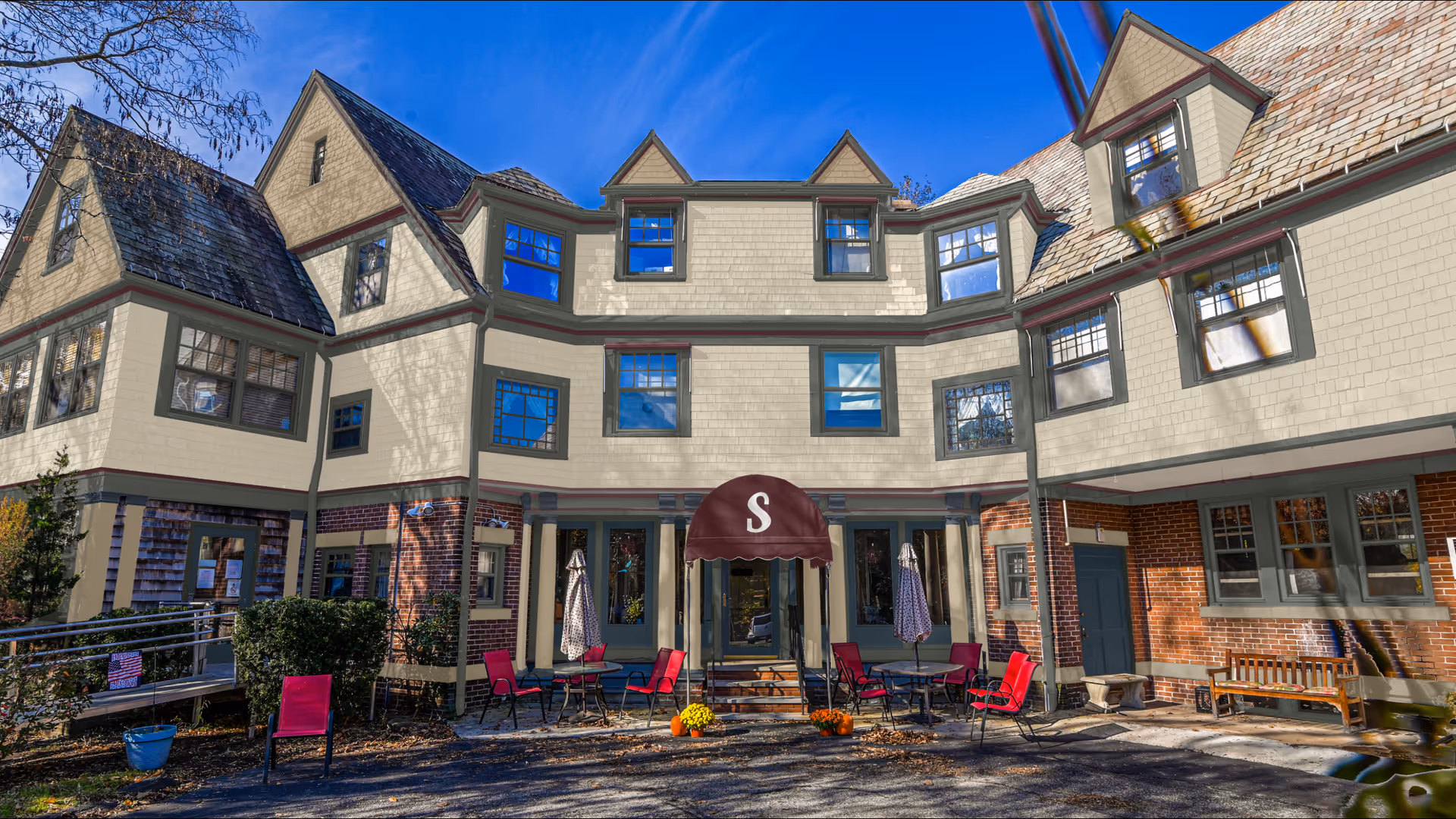 Exterior view of Sarawood Assisted Living facility showing a multi-story building with beige siding and brick accents. The building has multiple windows and a maroon awning with the letter 'S' above the entrance. There are outdoor tables and red chairs arranged in front of the entrance, along with some potted plants and a wooden bench on the right side.