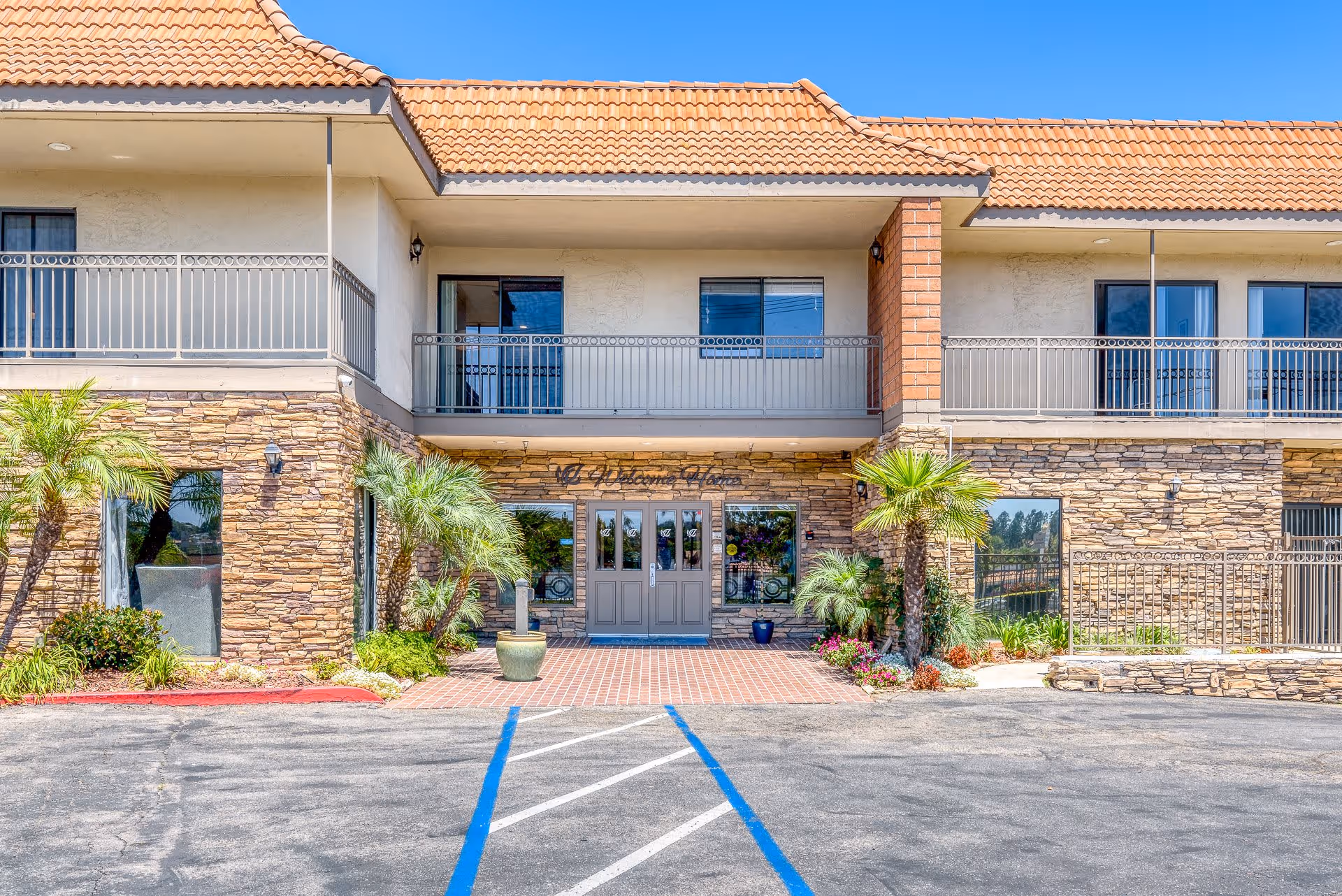 Front exterior view of Alta Vista Senior Living building with stone facade, beige walls, and a tiled roof. There are two palm trees and other plants near the entrance, which has double glass doors under a covered balcony with metal railings. The parking area in front has blue handicap parking lines.