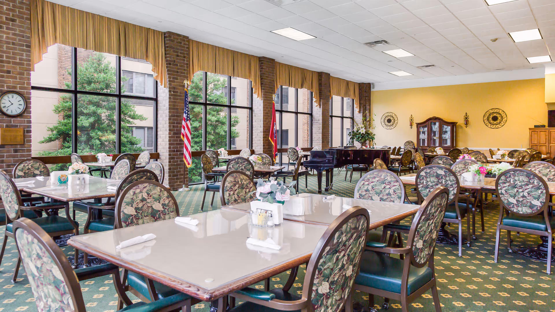 A spacious dining room with multiple tables and chairs arranged neatly. The chairs have floral patterned upholstery. Large windows with yellow curtains allow natural light to fill the room. There is a grand piano near the back wall, and American and Tennessee state flags are displayed near the windows. The room has a green carpet with a patterned design and yellow walls decorated with wall art and a wooden cabinet.